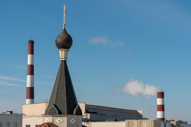 The dome with the cross of the Orthodox church on the background of the pipes of the chemical industry factory A church and a glass factory against a blue sky Faith in God and modern technology