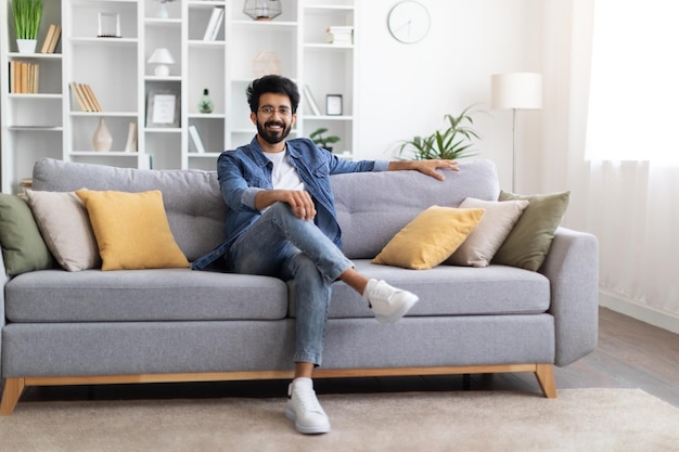 Домашний уют улыбающийся молодой индийский парень позирует на диване в гостиной