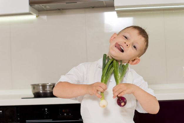 Domme jongen met tong die uitsteekt en groenten vasthoudt in de keuken