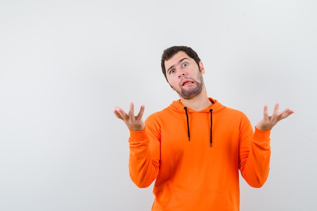 Domme knappe man in oranje hoodie handen opstijgen en op zoek dwaas geïsoleerd op witte achtergrond