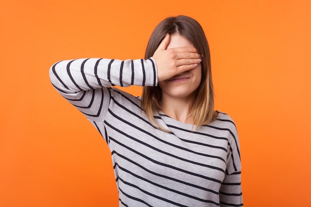 Don39t want to look at this Portrait of young scared woman with brown hair in long sleeve shirt standing covering eyes with hand refusing to watch indoor studio shot isolated on orange background