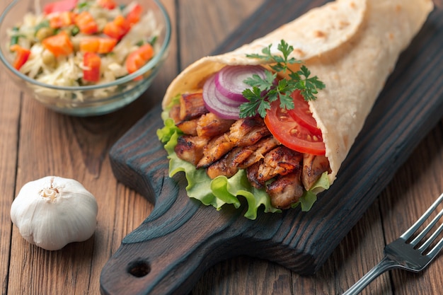 Doner kebab is lying on the cutting board. Shawarma with chicken meat, onions, salad lies on a dark old wooden table.