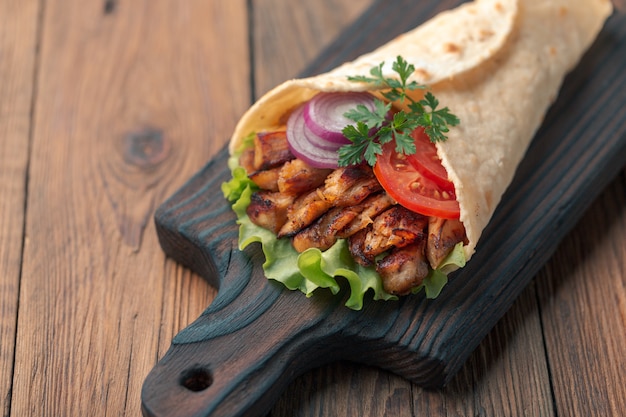 Doner kebab is lying on the cutting board. Shawarma with chicken meat, onions, salad lies on a dark old wooden table.