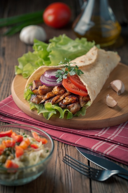 Doner kebab is lying on the cutting board. Shawarma with chicken meat, onions, salad lies on a dark old wooden table.