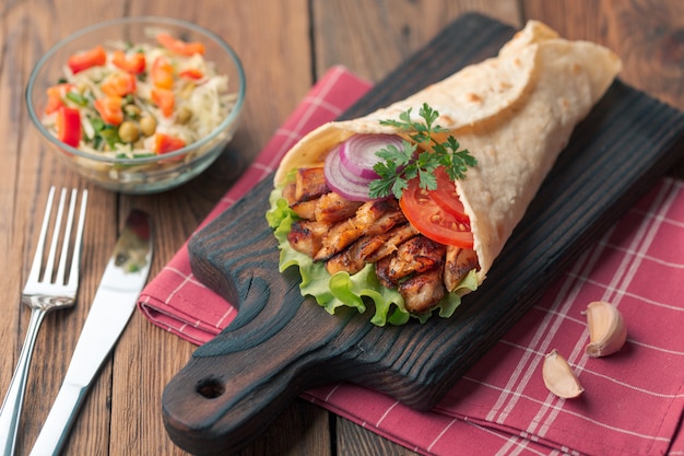 Doner kebab is lying on the cutting board. Shawarma with chicken meat, onions, salad lies on a dark old wooden table.