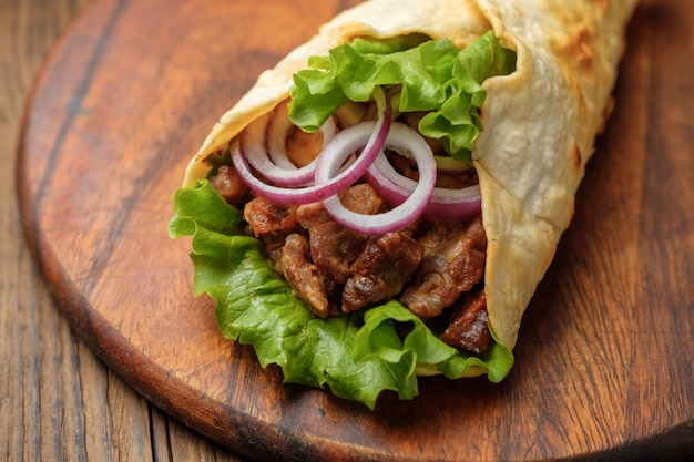 Doner kebab is lying on the cutting board. Shawarma with meat, onions, salad lies on a dark old wooden table.