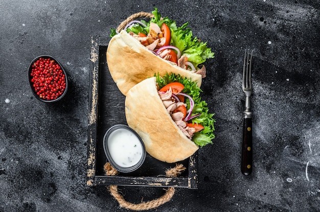 Doner kebab with grilled chicken meat and vegetables in pita bread on a wooden tray. Black background. Top view.