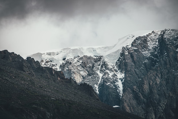 Donker atmosferisch surrealistisch landschap met donkere rotsachtige bergtop in lage wolken in grijze bewolkte hemel. Grijze lage wolk op hoge top. Hoge zwarte rots met sneeuw in lage wolken. Surrealistische sombere bergen.