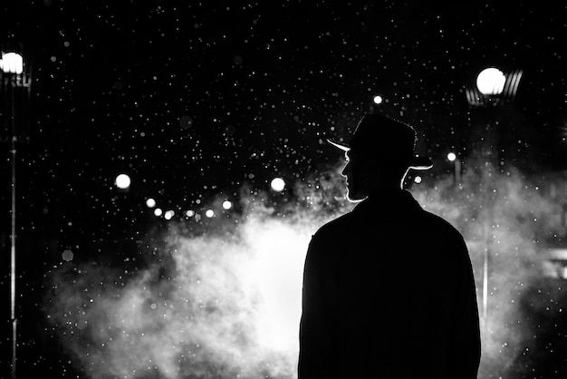 Donker silhouet van een man met een hoed in de regen op een avondstraat in een stad in de stijl van Noir