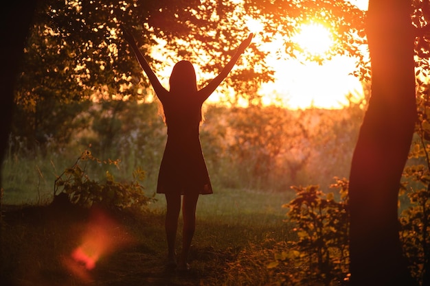 Donker silhouet van jonge vrouw die alleen door donker bos loopt en vreugdevol haar handen opsteekt in de zomeravond Genieten van het concept van natuur en buitenactiviteiten