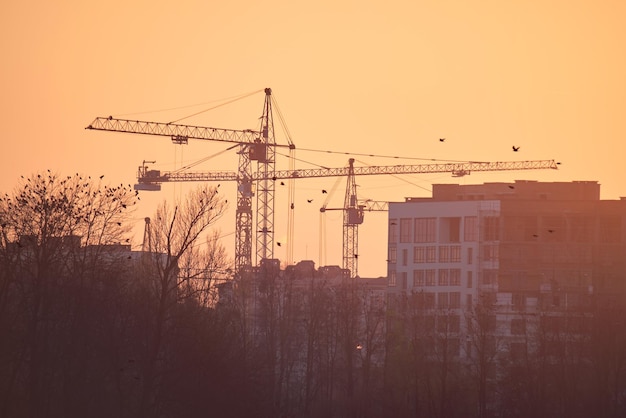 Donker silhouet van torenkranen op de bouwplaats van hoge residentiële flatgebouwen bij zonsondergang. Vastgoed Ontwikkeling.