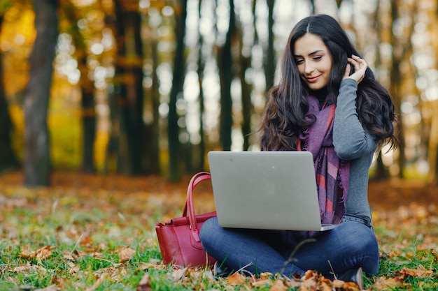 donkerbruin meisje dat aan haar laptop in het park tijdens de herfst werkt