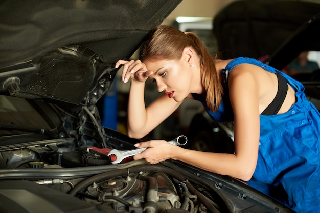 Donkerbruine meisjeswerktuigkundigen die of een auto herstellen inspecteren en een moersleutel in haar hand houden.