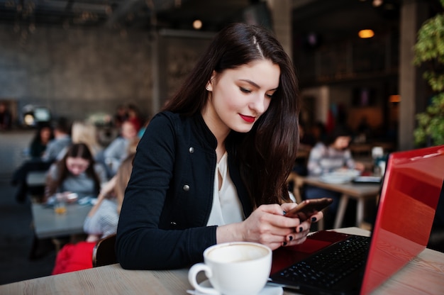 Donkerbruine meisjeszitting op koffie met kop die van cappuccino, met rode laptop werken en mobiele telefoon bekijken.