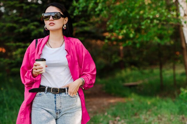 Donkerbruine mooie vrouw in zonnebril die koffiekop houdt en in het park loopt