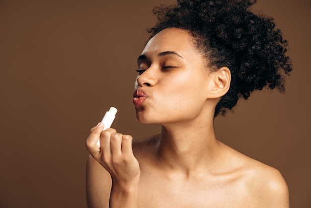 Donkerbruine multiraciale vrouw met schoonheidsgezicht met natuurlijke make-up en krullend haar die lippenbalsem op haar lippen toepassen. Lippenbeschermingsconcept