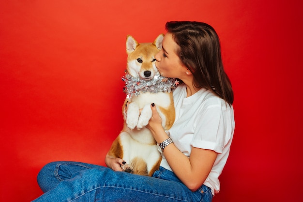 Donkerbruine vrouw die en Shiba Inu-hond omhelzen kussen in zilveren decoratie, rode achtergrond. Liefde voor de dieren