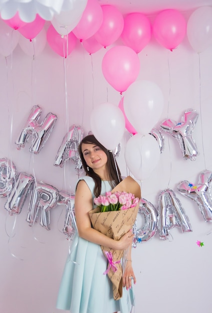 Donkerbruine vrouw in muntkleding die boeket van tulpen houdt. Verjaardagsversieringen met witte en roze kleur ballonnen, boeket tulpen en confetti voor feest op een witte muur. Gelukkige verjaardag verjaardag.