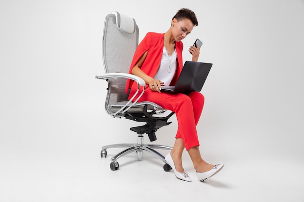 Donkerbruine zitting op stoel en het typen op laptop met een telefoon in haar handen op grijs