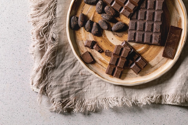 Donkere en melkchocoladereep heel en gehakt in keramische plaat met cacaobonen over grijze linnen doek als achtergrond Platte lay-ruimte