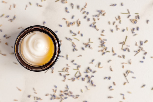 Donkere glazen pot met crème van lavendel, verspreide bloemen van gedroogde paarse lavendel bloesem op marmeren tafel.