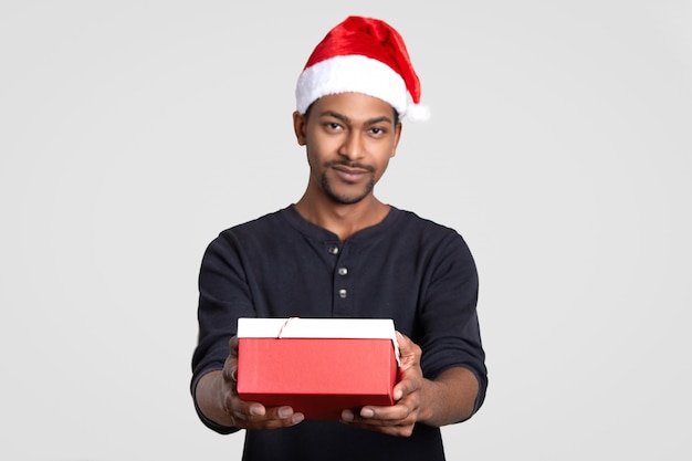 Donkere huid santa claus man houdt geschenkdoos, feliciteert u met nieuwjaar, modellen tegen witte muur. focus op het heden. etniciteit en feestdagen concept. african american man in feestelijke hoed