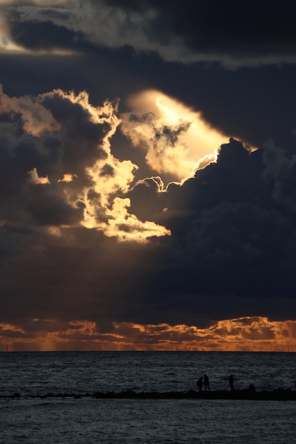 Donkere wolken met stralen tegen een zonsondergang boven de zee zwarte silhouetten van mensen aan de kust