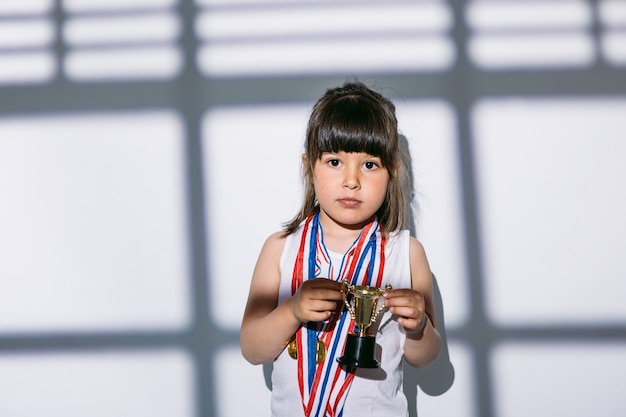 Donkerharig meisje met medailles voor sportkampioenschappen en trofeebeker, staande in de schaduw van een raam met een jaloezie over haar. sport en overwinningsconcept