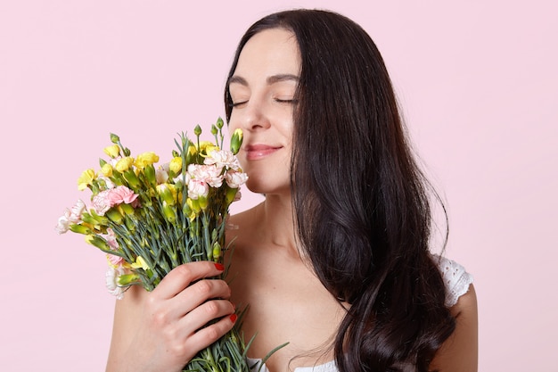 Donkerharige jonge dame, gekleed in witte jurk, heeft stijlvolle rode manicure, staat met gesloten ogen, ruikt aan mooie bloemen, kijkt zachtjes, houdt van haar boeket, brunette poseert over roze