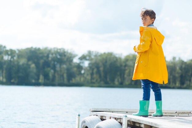 Donkerharige jongen die regenjas draagt die dichtbij rivier staat