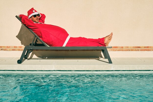 Donkerharige jongen in zwarte zonnebril verkleed als de kerstman liggend in een hangmat aan de rand van een zwembad in de zon