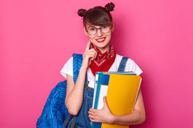 Donkerharige student met blauwe tas, houdt kleurrijke papieren map vast, glimlacht, houdt vinger op haar wang. Jong meisje draagt een t-shirt, denim overall met rode bandana op de nek. Tiener komt terug naar school.