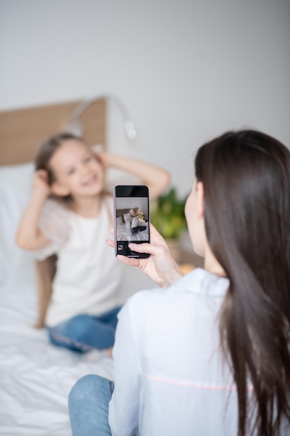 Donkerharige vrouw zittend op het bed die een glimlachend vrolijk meisje fotografeert met de smartphone