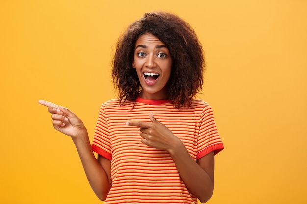 donkerhuidig jong meisje met afrokapsel in gestreepte t-shirt wijzend naar links opgetogen en gefascineerd poseren tegen oranje muur