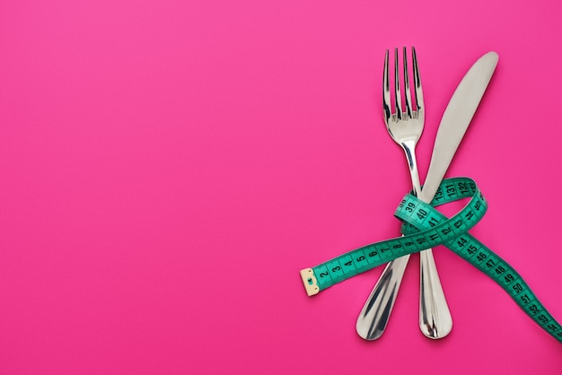 Dont eat too much. Knife and fork crossed, connected by measure tape isolated on pink background