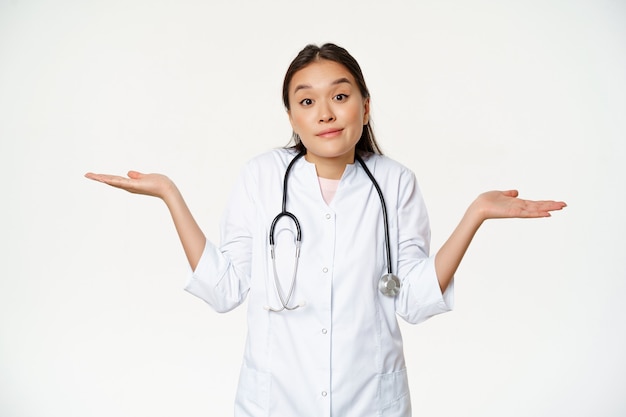 Dont know. Clueless female asian doctor shrugging, spread hands sideways and looking puzzled, cant understand, standing over white background