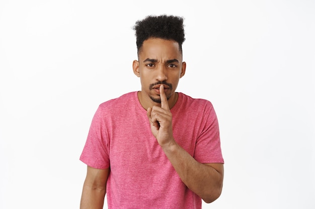 Dont speak. Serious african american guy hushing, hold finger near lips while frowning upset, shushing, tell to be quiet, scolding loud person, white background, pink t-shirt