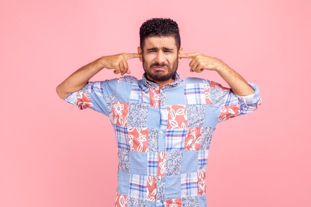 Dont want to listen Portrait of bearded man wearing casual style blue shirt irritated by loud noise covering ears and grimacing in pain Indoor studio shot isolated on pink background