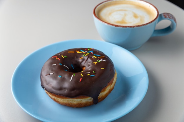 Donut covered with chocolate glaze and multicolored sprinkles on blue plate and cup of cappuccino