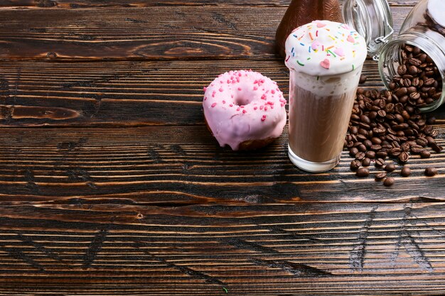 A donut with pink icing and chocolate powder and a cappuccino glass with high foam and decoration. A can of coffee and pouring grains.