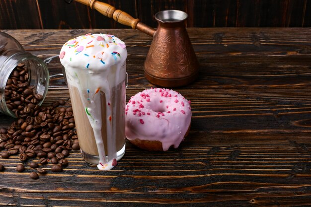 A donut with pink icing and chocolate powder and a cappuccino glass with high foam and decoration. A can of coffee and pouring grains.