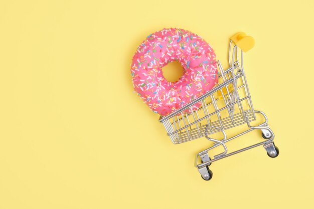 Donut with sprinkles and pink icing in a shopping cart on yellow surface top view copy space