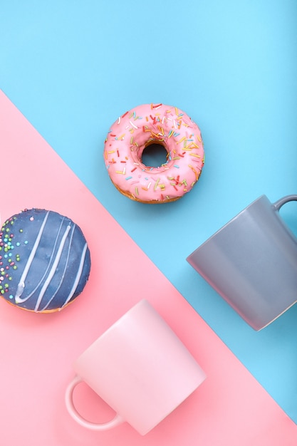 Donuts met glazuur en koffiekopjes op pastelblauw en roze oppervlak, zoete donuts.