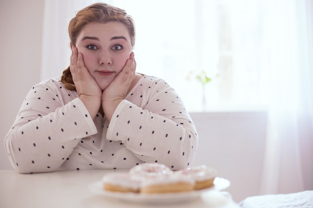 Пончики на тарелке. Озадаченная пухлая женщина сдерживается, сидя рядом с вкусной тарелкой пончиков