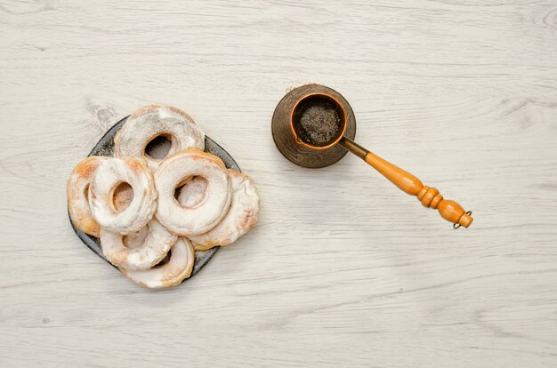 Donuts sprinkled with powdered sugar, fresh coffee in cezve, light wooden background. Top view