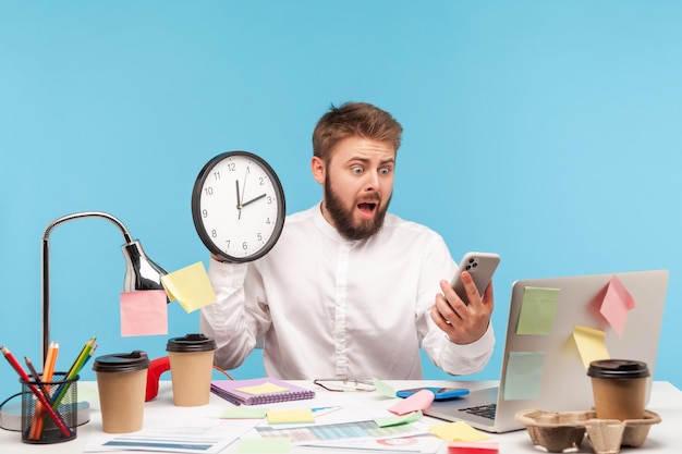 Foto doodsbang nerveuze man kantoormedewerker wandklok te houden en te kijken naar smartphone-display met angst laat op afspraak ontbrekende zakelijke bijeenkomst indoor studio shot geïsoleerd op blauwe achtergrond