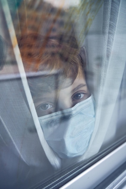 Door glas zicht op een preteen-kind dat een beschermend medisch masker draagt en naar de camera kijkt terwijl hij achter een raam met gordijnen staat tijdens een pandemie van het coronavirus