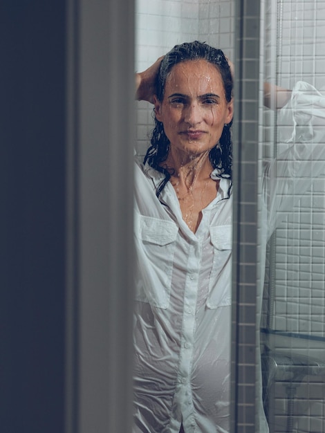 Door glazen deur aantrekkelijke verleidelijke vrouw in wit nat overhemd hoofd aanraken en naar de camera kijken terwijl ze onder waterdruppels in de douchecabine staat