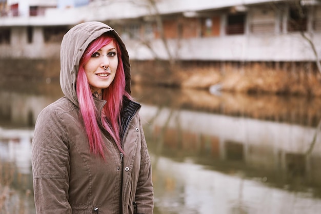 Foto doorboorde jonge vrouw met roze haar en parka met capuchon die bij de rivier staat