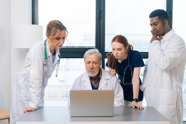 Doordacht medisch team van multi-etnische artsen die samen praten op zoek naar een laptopscherm dat op de achtergrond van het raam staat